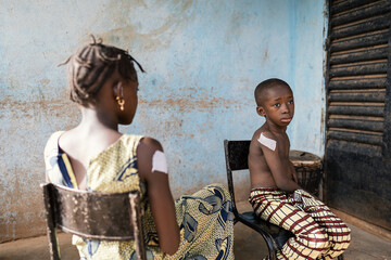 Two black siblings sitting outside a rural dispensary with big plasters on their upper arms after a...