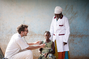 Reassuring white doctor auscultating the chest of a serious little African girl with a black nurse...