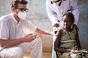 White doctor preparing a vaccine shot to a cute small black schoolgirl assisted by a local health...