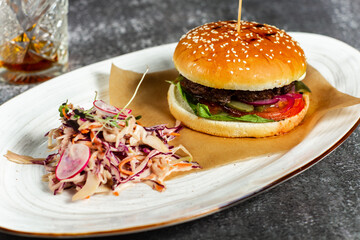 Fresh tasty burger with soft bun meat cutlet and fresh vegetables served with vegetable salad, White oval plate with whiskey on background. Horizontal orientation