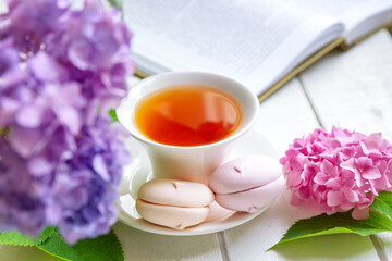 A cup of tea surrounded by hydrangea flowers, with pink marshmallows and an open book. Summer mood, tea party concept.