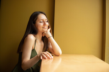 beautiful girl in summer dress dreams of closing her eyes while sitting at the table in cafe inside against a yellow wall