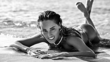 Sexy slim woman girl in bikini is lying on the sand of the beach in the incoming waves of the sea during vacation on seashore, getting tan, holding feet up. Black and white. Summer vibes