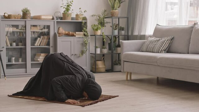 Slowmo shot of young Muslim woman in traditional black dress and hijab praying on floor at home
