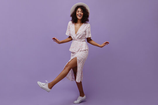 Joyful dark-skinned lady in white polka dot dress smiles widely on purple background. Happy curly woman in hat moves on isolated.