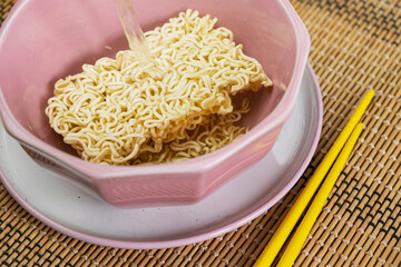 Quick instant dry noodles with pouring hot water in pink bowl on bamboo mat, Asian Chinese ramen pasta fast food eaten with yellow bamboo chopsticks