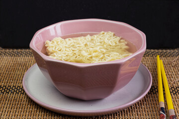 Quick instant dry noodles with hot water in pink bowl on black background, Asian Chinese ramen pasta fast food eaten with yellow bamboo chopsticks