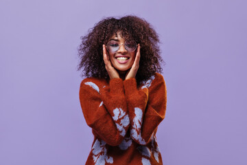 Portrait of curly brunette woman in eyeglasses looking into camera on purple background. Happy girl in warm burgundy sweater smiles on isolated.
