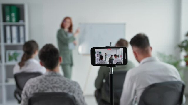 online training, a young female entrepreneur makes a presentation to a working team showing a graph on a flipchart during a video broadcast using a smartphone in an office