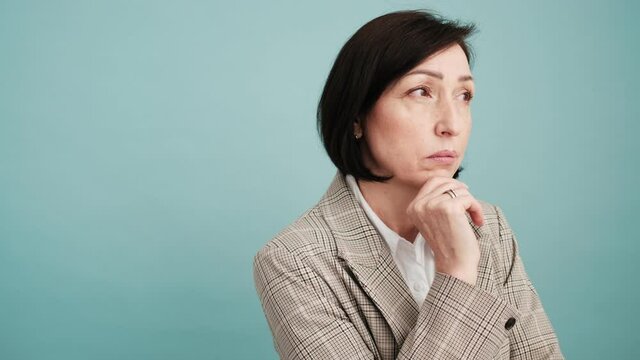 A concentrated mature woman is thinking about something while looking to the side standing isolated over blue wall in the studio