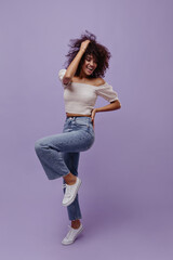 Excited brunette curly woman in jeans and cropped white top moves, smiles, touches hair and rises leg on purple background.