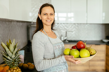 Portrait of mature brunette caucasian woman housewife in apron holding plate of fruit kitchen woman in kitchen at home. Healthy domestic lifestyle concept.