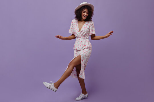 Good-humored brunette woman in white dress and hat moves on purple background. Attractive dark-skinned lady smiles.