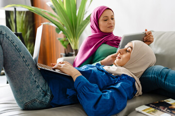Two women using tablet computer and laptop while resting on sofa