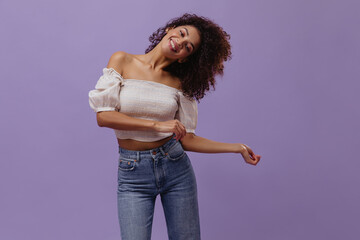 Pretty dark-skinned woman in jeans abs white top plays hair on purple background. Curly brunette...
