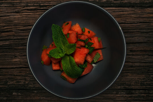 Melon pieces served with green mint leaves in a grey bowl on dark brown wooden table