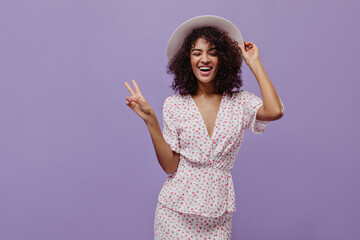 Joyful brunette woman in stylish polka-dot dress winks and shows peace sign on purple background....