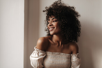 Attractive dark-skinned woman leans on white wall. Pretty curly brunette lady in stylish top smiles...