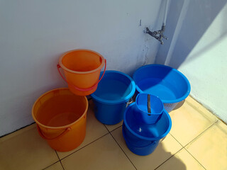 high angle of blue and orange plastic bucket which filled with water near the water tap on the courtyard of the asian urban house with light brown tiles and white walls in the morning with a sunlight