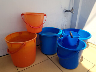 The blue and orange plastic bucket which filled with water near the water tap on the courtyard of the building with light brown ceramic floor tiles and white walls in the morning with a sunlight © yayang