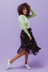 Curly brunette woman in midi floral dress and green top dances on purple background. Happy...