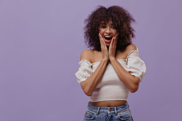 Surprised curly woman looks into camera on isolated. Happy brunette dark-skinned lady in white top poses on purple background.