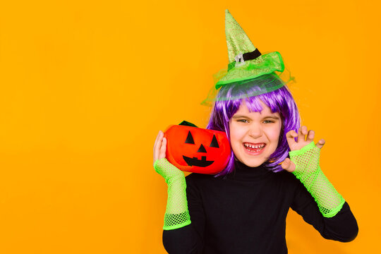 Scary girl kid in a witch costume with a green hat holds a pumpkin face. Halloween, traditions, horror stories concept.