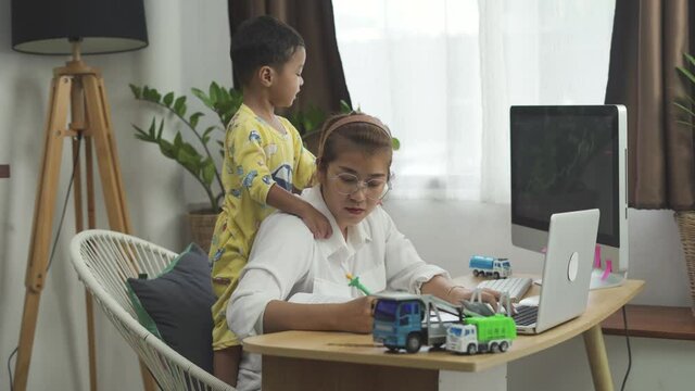 A Busy Young Asian Mother Tries To Work Remotely With Her Child At Home. The Boy Interferes With The Mother's Work. 