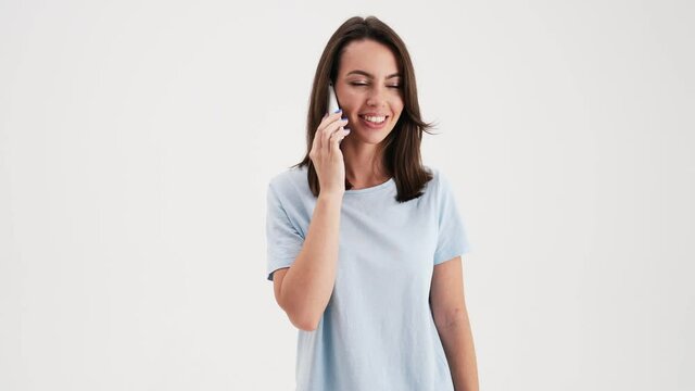 A happy woman wearing basic t-shirt is talking on her mobile standing in the studio isolated over white background