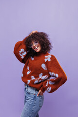 Curly dark-skinned woman in warm sweater and jeans touches hair. Attractive lady in eyeglasses smiles on purple background.