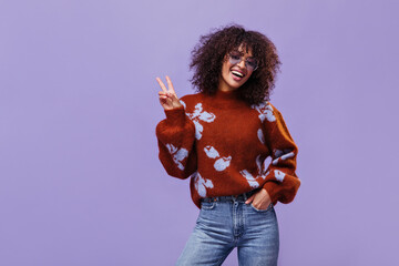 Curly brunette dark-skinned woman smiles sincerely and shows peace sign. Joyful girl poses on...
