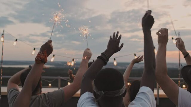 Handheld shot of group of young people with sparklers partying on rooftop terrace while DJ playing set on summer evening