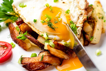 Fried potatoes with egg with liquid flowing yolk, herbs and tomatoes on a white plate.
