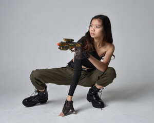 Full length portrait of pretty brunette, asian girl wearing black top and khaki utilitarian army pants and leather boots. Sitting pose holding a science fiction gun, isolated agent a light grey studio