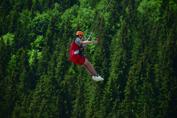 A young man jumped from bungee jumping and now hangs on a rope and films himself on a sports video camera against a blurred background of a green forest