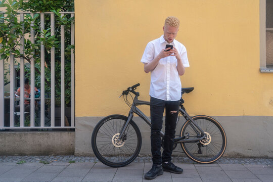 Germany, Cologne, Albino man using smart phone in front of building and bicycle - Powered by Adobe