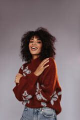 Brown-eyed brunette woman in warm burgundy sweater smiles on grey background. Curly lady with beautiful makeup poses on isolated.