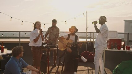 Handheld tracking shot of band playing music at party on rooftop terrace on windy summer evening