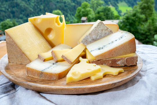 Cheese collection, wooden board with French cheeses comte, beaufort, abondance, emmental, morbier and french mountains village in Haute-Savoie on background