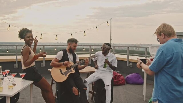 Handheld shot of young man with mobile phone filming his friends dancing and playing song on acoustic guitar on rooftop terrace