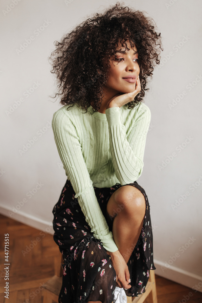 Wall mural Dark-skinned brunette woman in black floral skirt and green top looks away and smiles. Charming lady sits on chair near white wall.