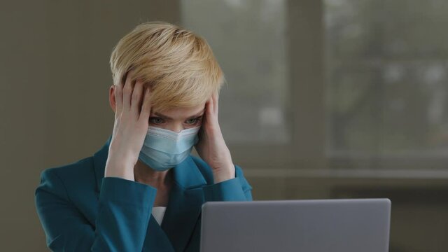 Portrait of mature blonde middle-aged business woman wears protective medical mask works with laptop receives bad news letter refusal from bank holds head with hands from headache problem pressure