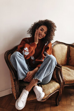 Cute brunette woman in denim pants and burgundy sweater smiles and sits on classic style armchair in white room.