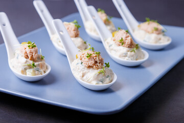 Cold appetizer made from cod liver, cod caviar, olives, cucumber and microgreens on serving spoons. Traditional cold dish. Close-up, blue plate, black background.