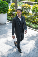 Successful man in formal black suit and sunglasses walking on the street and looking aside.
