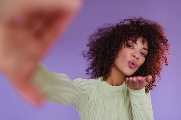 Brunette curly woman blows kiss on purple background. Attractive dark-skinned lady in green top...