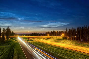 Fototapeta premium Motion blur of busy highway going through rural landscape with nice clouds at night and car light trails with long exposure. Background 