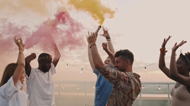 Handheld shot of group of happy young people holding color smoke bombs and partying together on rooftop terrace on summer evening