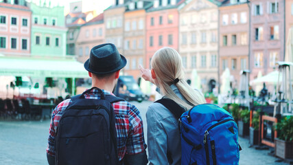 Back view of young tourists couple with bags checking map on central city square. They discussing their new destination.