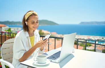 Young elegant woman wearing wireless headphone looking at smartphone with smile while sitting at balcony with seaview on resort. girl watching video, chatting in social network - 447728532